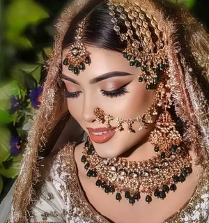 Bride in a garden setting with dark green and gold jewelry, eyes closed in a serene expression.