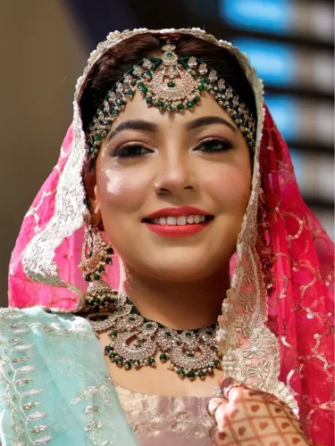 Smiling South Asian bride in pink attire with detailed headpiece and makeup.
